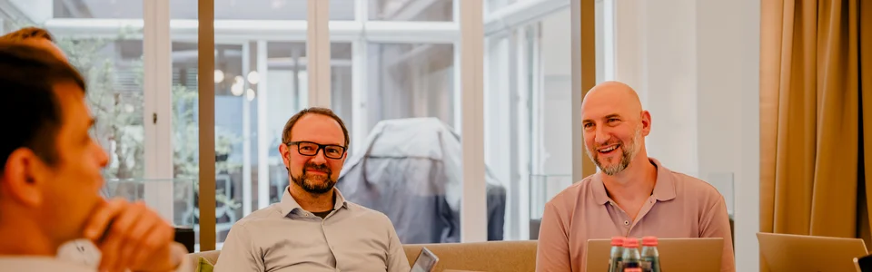 Mehrere Personen bei einem Meeting; Laptops und Wasserflaschen auf dem Tisch; moderner Konferenzraum.