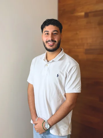 Eine Person mit weißem Polohemd steht vor einer Wand, lächelt und hat die Hände vor sich verschränkt.