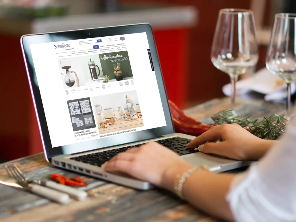Person am Laptop beim Online-Shopping auf einer Küchengeräte-Website. Im Hintergrund stehen zwei Weingläser auf einem Tisch.