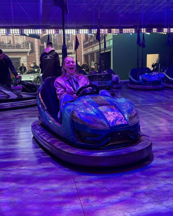 Person fährt in einem lila Glitzer-Autoscooter auf einer Indoorbahn, umgeben von weiteren Fahrern unter leuchtenden Lichtern.