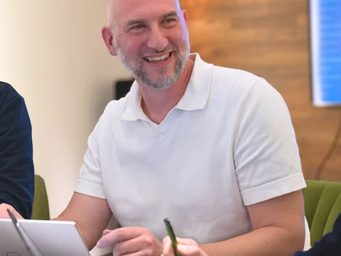 Ein Mann mit Glatze lächelt bei einem Treffen in einem modernen Büro, umgeben von Laptops und blauer Präsentationsfolie.