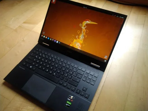 Laptop auf Holzfußboden, geöffneter Bildschirm mit orangefarbenem Hintergrund und abstraktem Design, Tastatur sichtbar.