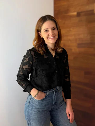 Eine Frau steht lächelnd in einem Raum mit Holzwand, trägt eine schwarze Bluse und blaue Jeans.
