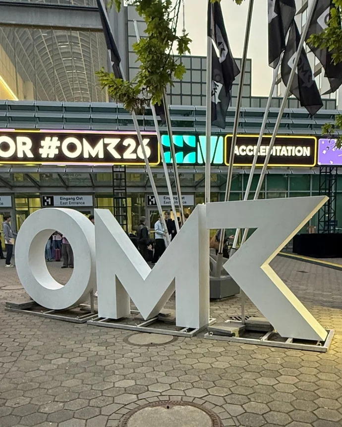 Große "OMK" Buchstaben vor einem Eingang mit Bannern, aufgenommen bei einer Messe oder Konferenzveranstaltung.