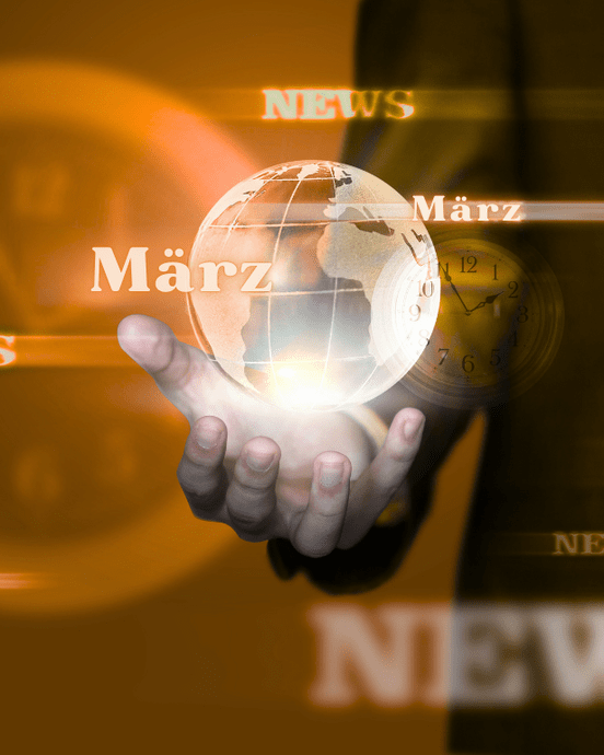 Hand hält rotierende Weltkugel mit Text "März" und "News" vor verschwommenem Uhr-Hintergrund in warmen Brauntönen.