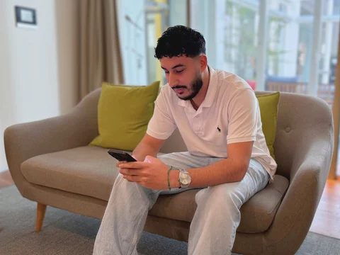 Mann sitzt auf einem Sofa und schaut auf sein Smartphone. Im Hintergrund sind helle Fenster und grüne Kissen zu sehen.