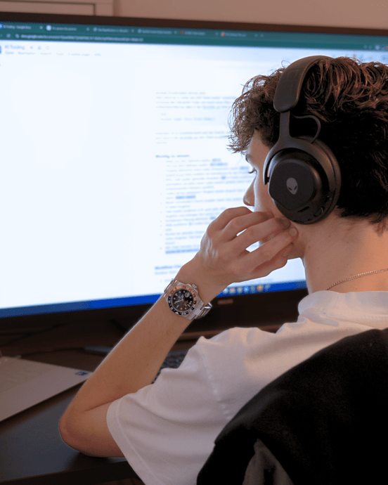 Person mit Kopfhörern arbeitet an zwei Bildschirmen, ein Laptop mit Codierung, ein großer Bildschirm zeigt ein Dokument.