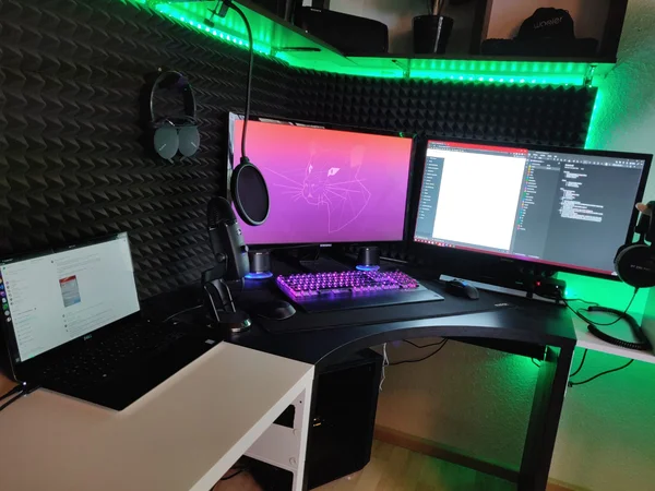Moderne Gaming- und Arbeitsstation mit zwei Monitoren, Laptop, RGB-Tastatur und Tonaufnahme-Equipment, umgeben von Schallschutzmatten.