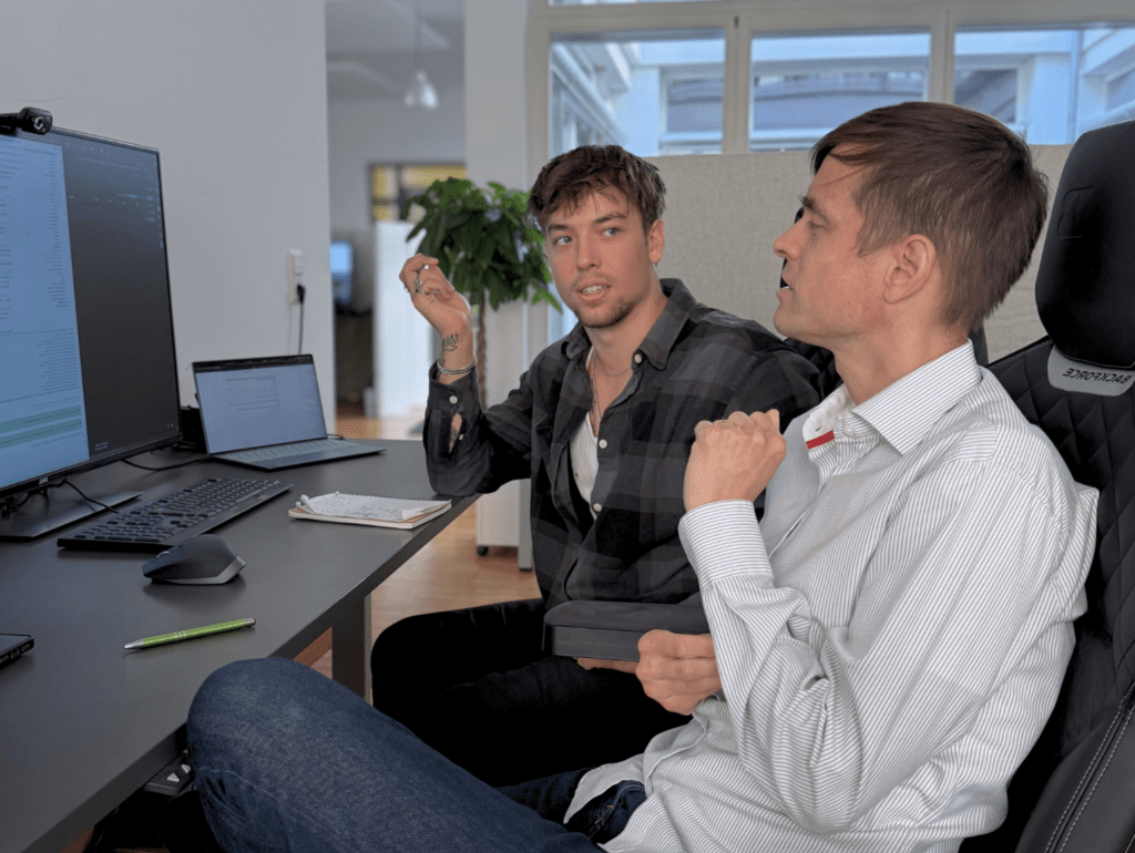 Zwei Männer diskutieren vor Computermonitoren in einem modernen Büro, umgeben von Dokumenten und Kaffeetassen.