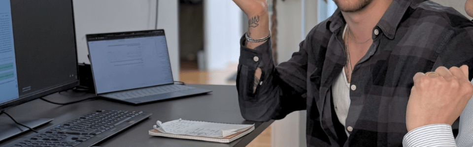 Zwei Männer diskutieren vor Computermonitoren in einem modernen Büro, umgeben von Dokumenten und Kaffeetassen.