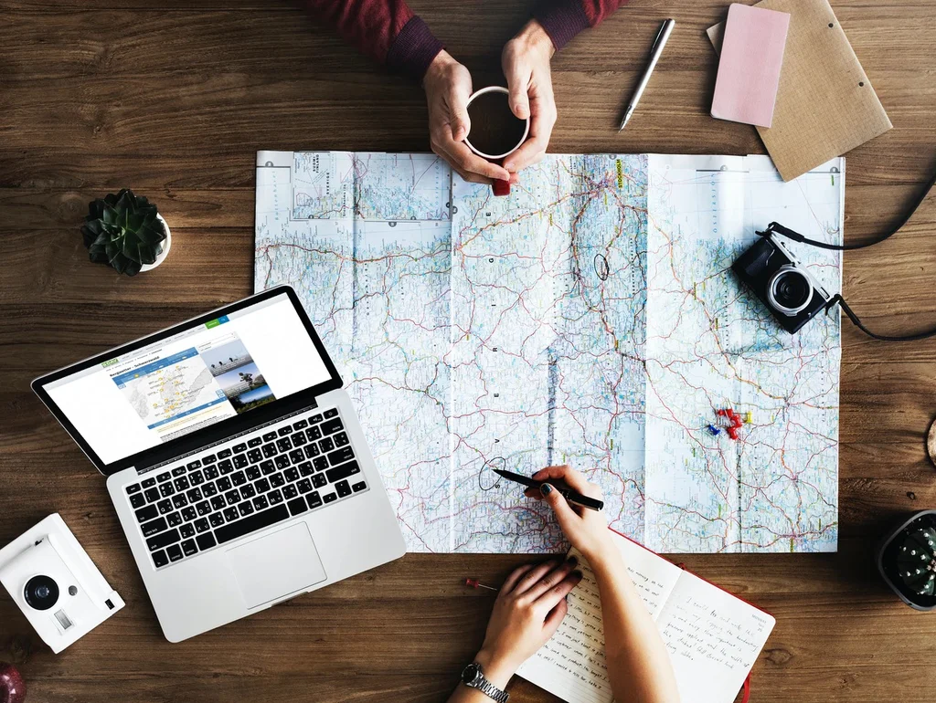 Personen planen eine Reise; Karten, Laptop, Kamera und Notizen auf Holztisch. Hände halten Tasse und Stift.