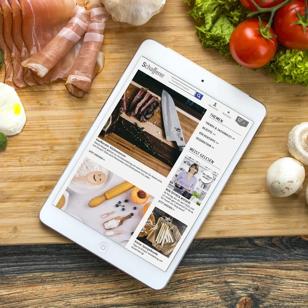 Tablet mit Rezeptwebsite auf Holzbrett, umgeben von Zutaten wie Tomaten, Pilzen, Schinken, Basilikum und Mozzarella.