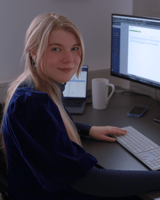 Person arbeitet an einem großen Monitor und Laptop, umgeben von Tastatur, Smartphone und Tasse auf einem Schreibtisch.