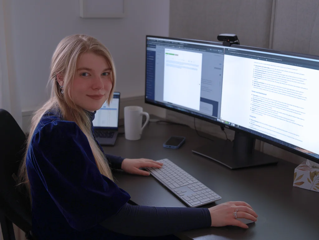 Person arbeitet an einem großen Monitor und Laptop, umgeben von Tastatur, Smartphone und Tasse auf einem Schreibtisch.