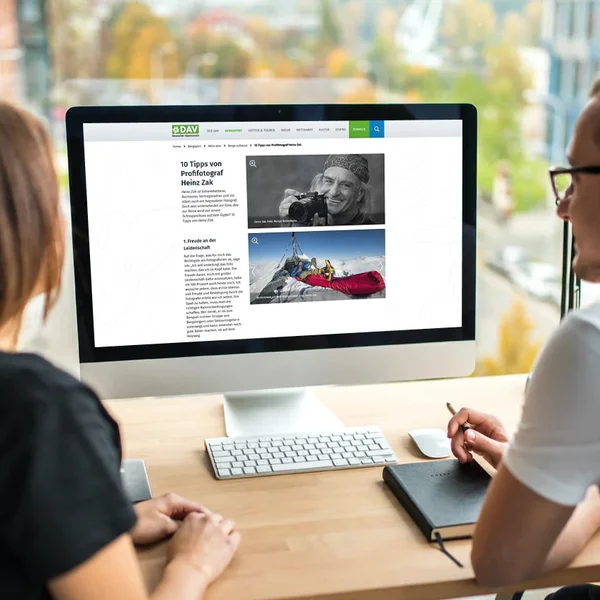 Zwei Personen betrachten einen Computerbildschirm mit einem Artikel über Fotografie in einem modernen Büro mit Stadtblick.