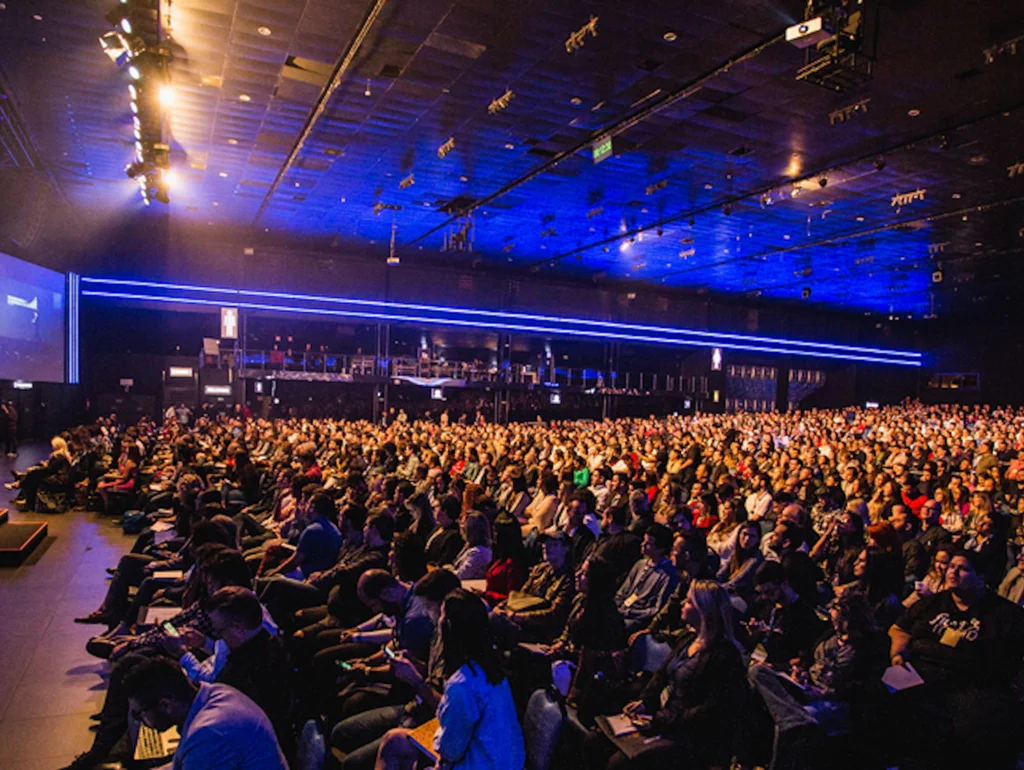 Großer Konferenzsaal mit Publikum vor einer Bühne und Leinwand, beleuchtet mit blauem Licht, Präsentation im Gange.