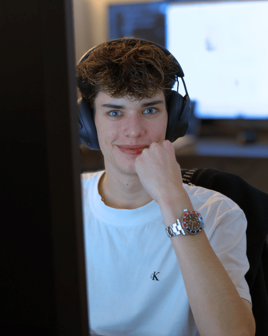 Ein Mann mit Kopfhörern sitzt am Computer und schaut lächelnd auf den Bildschirm, ein Monitor im Hintergrund.