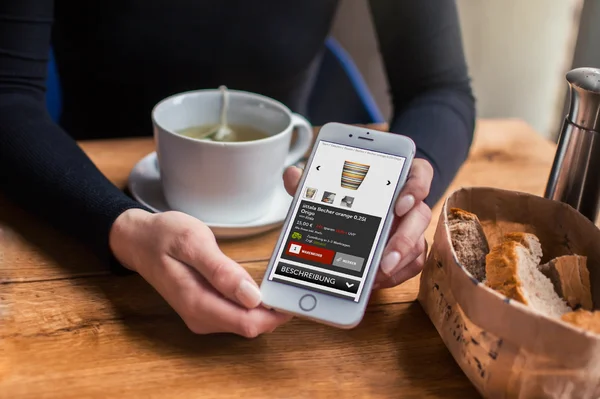 Person hält Smartphone mit Onlineshop; im Hintergrund eine Tasse Tee und ein Brotkorb auf Holztisch.