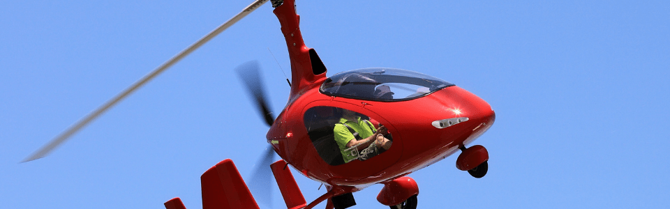 Roter Tragschrauber fliegt unter klarem Himmel, Pilot im Cockpit sichtbar.