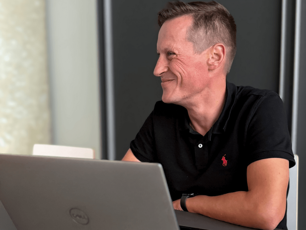 Eine Person sitzt lächelnd an einem Tisch mit einem geöffneten Laptop in einem Büro.