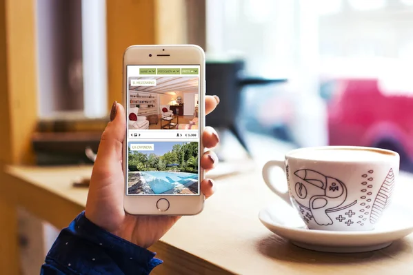 Smartphone zeigt Ferienunterkünfte, gehalten von Person in Café, daneben eine dekorierte Tasse.