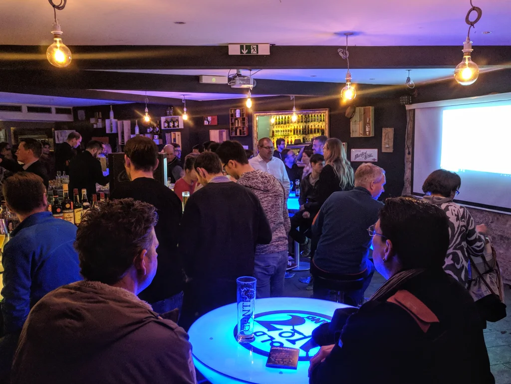 Menschen in einer beleuchteten Bar, die sich unterhalten; Sitzgelegenheiten und eine Leinwand sind sichtbar.