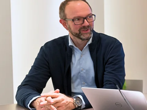 Ein Mann mit Brille sitzt an einem Tisch mit Laptop und Notizblock, lächelnd in einem hell beleuchteten Raum.