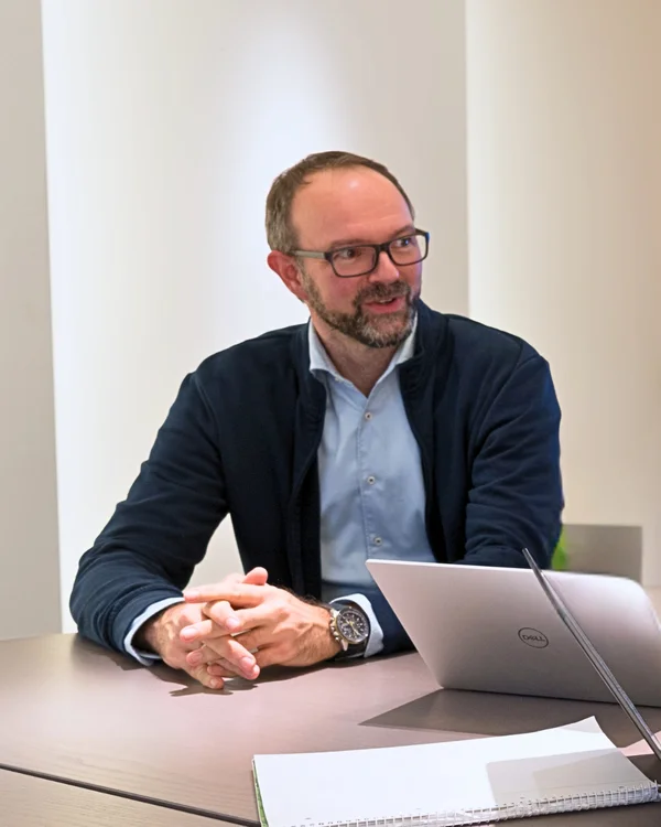 Ein Mann mit Brille sitzt an einem Tisch mit Laptop und Notizblock, lächelnd in einem hell beleuchteten Raum.