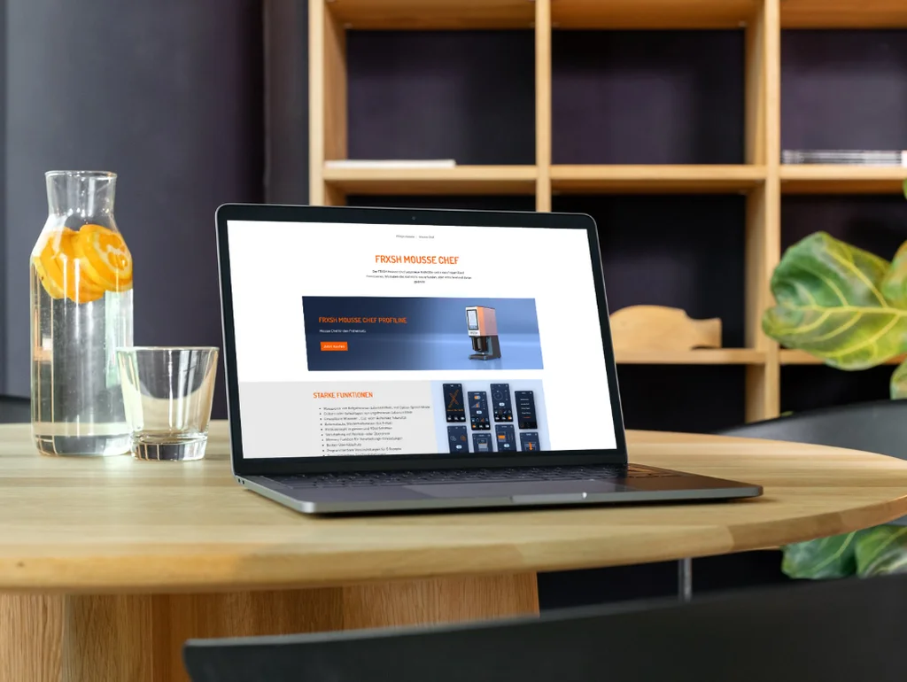 Laptop auf Holztisch mit geöffneter Webseite, flankiert von einer Karaffe mit Wasser und einem Glas, im Hintergrund Regale.
