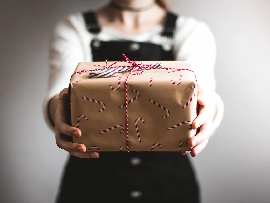 Person überreicht Geschenk in braunem Papier mit Zuckerstangendruck, rosa-weißem Band, unscharfer Hintergrund.