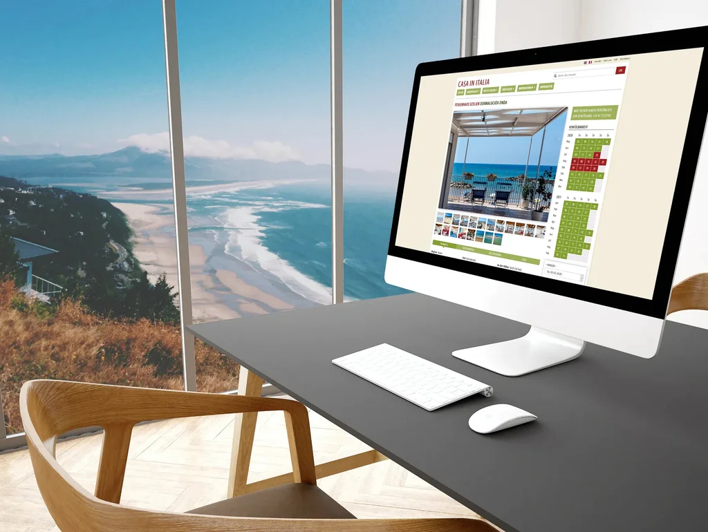 Computer auf Schreibtisch mit Blick auf eine Küstenlandschaft durch Fenster; Bildschirm zeigt eine Immobilienseite.