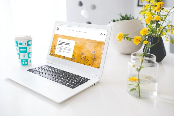 Laptop auf weißem Schreibtisch mit Blumen und Kaffeebecher, zeigt eine Website mit gelbem Hintergrund.