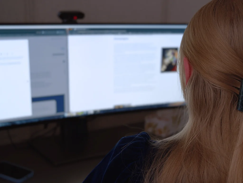 Person arbeitet an einem Computer mit zwei Bildschirmen, liest Textinhalt.