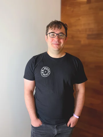 Ein Mann mit Brille trägt ein schwarzes T-Shirt vor einer teils weißen, teils holzverkleideten Wand.