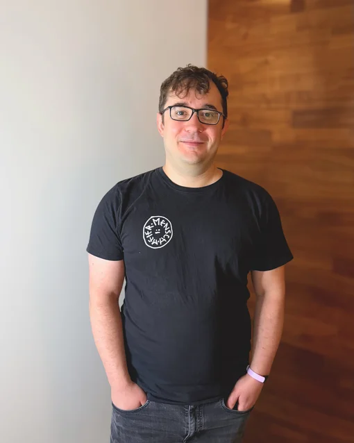 Ein Mann mit Brille trägt ein schwarzes T-Shirt vor einer teils weißen, teils holzverkleideten Wand.