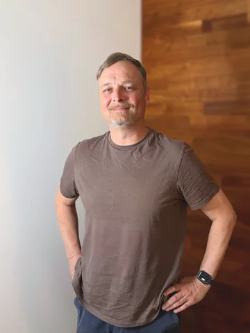 Eine Person im braunen T-Shirt steht lächelnd vor einer zweifarbigen Wand.