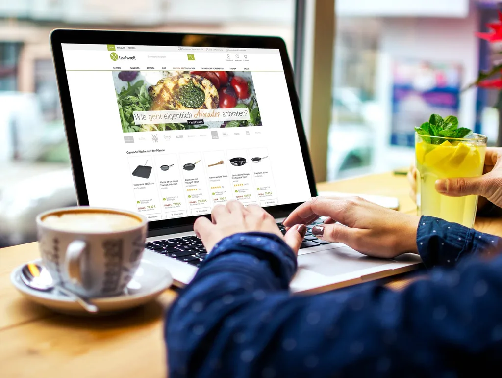 Person tippt auf Laptop in Café, Webseite mit Küchengeräten geöffnet; daneben Kaffee und Limonade mit Minze.