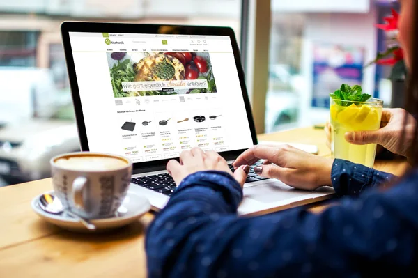 Person tippt auf Laptop in Café, Webseite mit Küchengeräten geöffnet; daneben Kaffee und Limonade mit Minze.