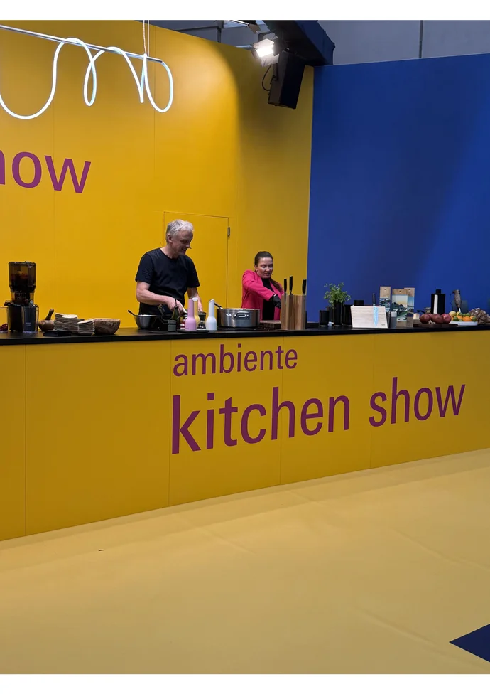 Zwei Personen präsentieren auf einer gelben Bühne der "ambiente kitchen show" moderne Küchengeräte.