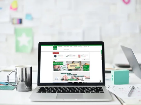 Laptop auf Schreibtisch vor weißer Wand, geöffnet mit Webseite auf dem Bildschirm; daneben Tasse und Dokumente.