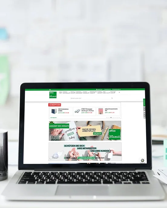 Laptop auf Schreibtisch vor weißer Wand, geöffnet mit Webseite auf dem Bildschirm; daneben Tasse und Dokumente.