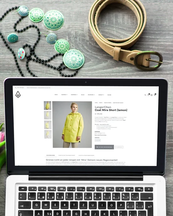 Laptop mit Online-Shop-Seite, umgeben von rosa Tulpen, Kaffee, Accessoires und Kleidung auf Holzuntergrund.