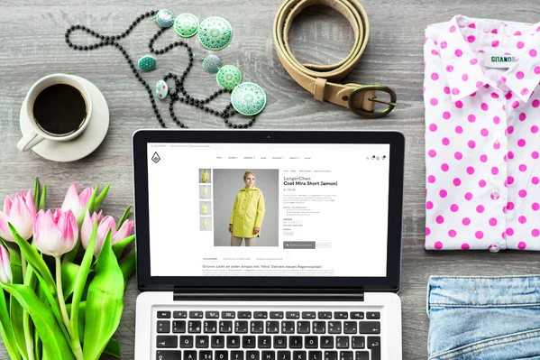 Laptop mit Online-Shop-Seite, umgeben von rosa Tulpen, Kaffee, Accessoires und Kleidung auf Holzuntergrund.