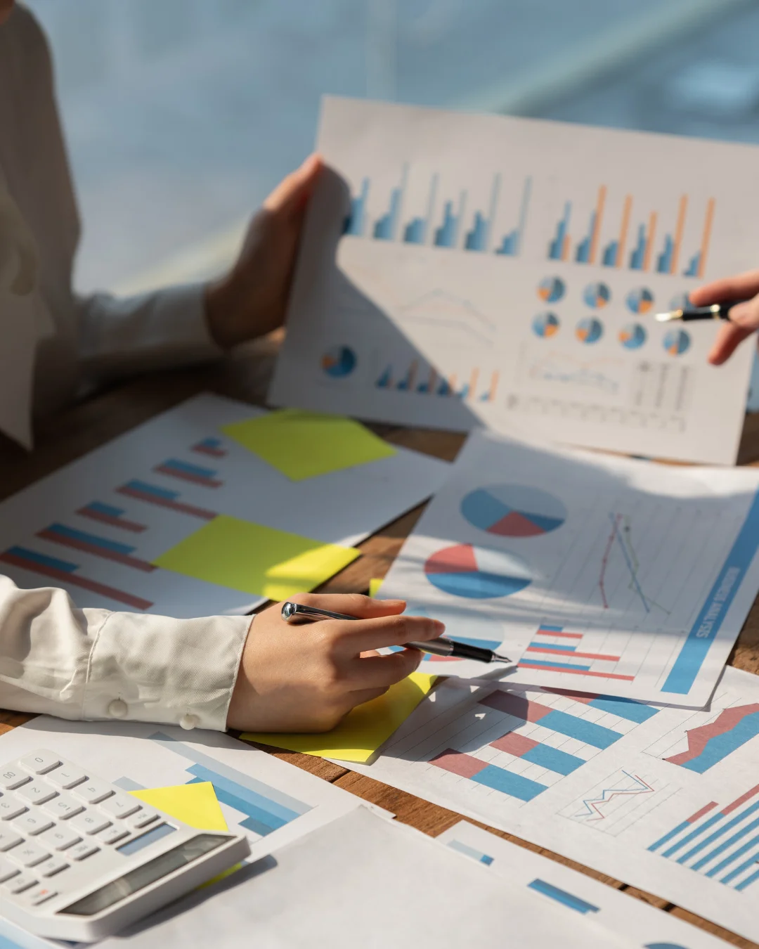 Personen analysieren Finanzdiagramme auf Schreibtisch. Tabellen, Diagramme und ein Taschenrechner sind sichtbar.