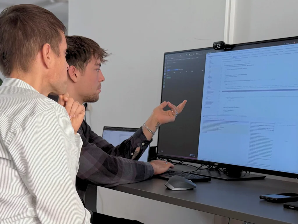 Zwei Männer arbeiten an einem großen Monitor mit Code-Editor. Einer zeigt auf den Bildschirm. Im Hintergrund steht ein Laptop.