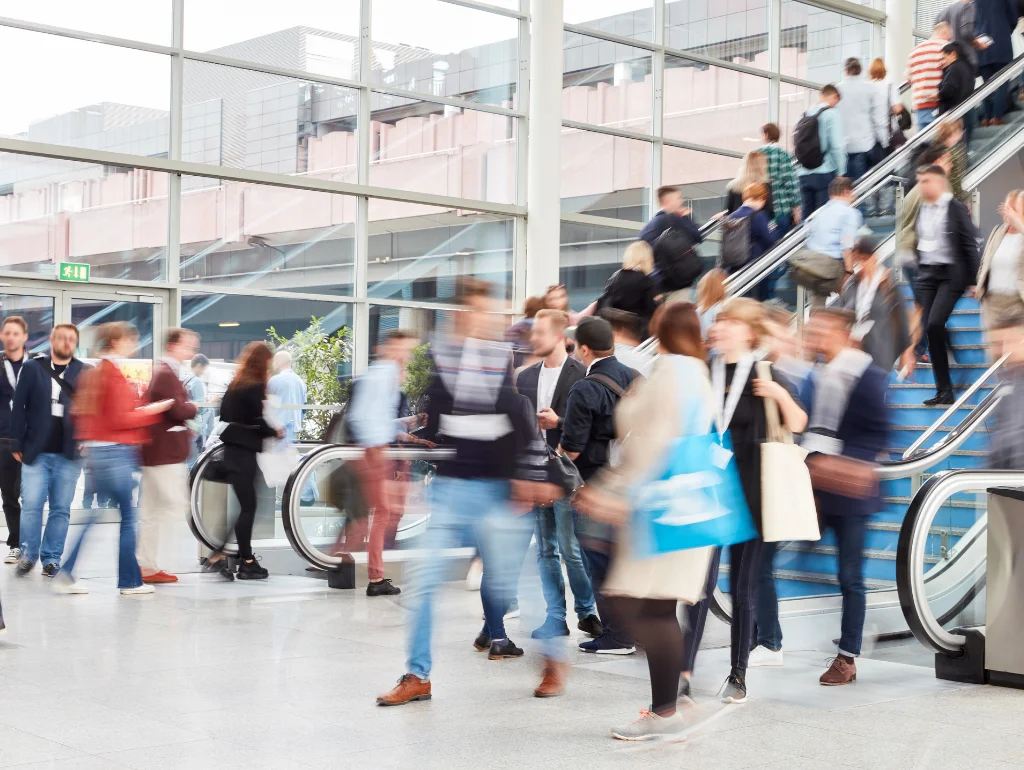 Menschenmenge in Bewegung auf einer Messe, einige auf Rolltreppen, moderne Architektur im Hintergrund.