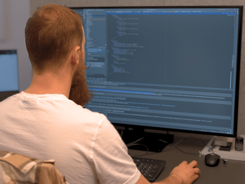Mann arbeitet an einem Computer mit großem Monitor, Programmiercode auf dem Bildschirm sichtbar.