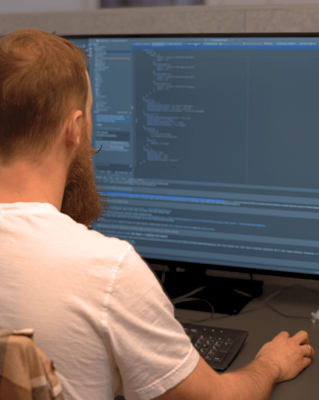 Mann arbeitet an einem Computer mit großem Monitor, Programmiercode auf dem Bildschirm sichtbar.
